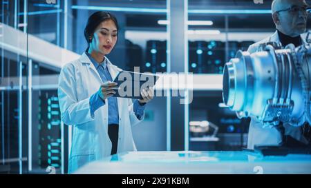 Middle Aged Engineer and Young Asian Scientist Collaborate on a New Type of Electric Turbine Engine, Standing and Using a Tablet Computers in Scientific Technology Laboratory. Stock Photo