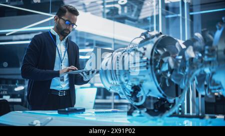 Chief Engineer Using Laptop Computer, Analyzing and Researching How a Futuristic Turbofan Motor Works. Senior Manager Developing Innovative Technology in Industrial High Tech Facility. Stock Photo