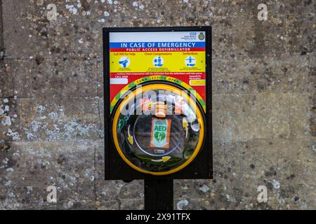 Public access defibrillator in a yellow case mounted on poll in front of a  textured wall with emergency instructions. Stock Photo