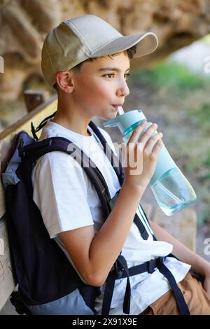 preteen boy hiking in the mountains with cap and backpack drinking water sitting on a bench Stock Photo