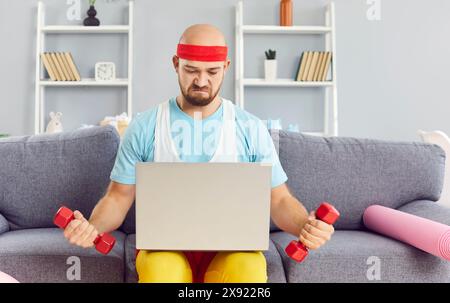 Funny man using dumbbells exercising following trainer online via laptop sitting on sofa at home. Stock Photo