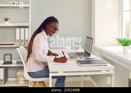 African American woman accountant sitting at office desk, working on laptop and taking notes Stock Photo