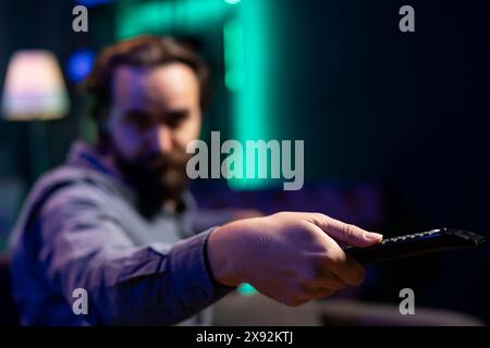 Focus on remote control used by man in blurry background switching channels on television screen. Close up of device used by person to scroll through streaming content, picking movie Stock Photo