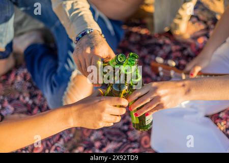 closeup shot captures the hands of three Latin friends raising beer bottles in a toast on the beach during a summer sunset. The image portrays the cam Stock Photo