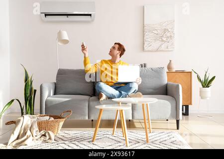 Young man with laptop switching on air conditioner in living room Stock Photo