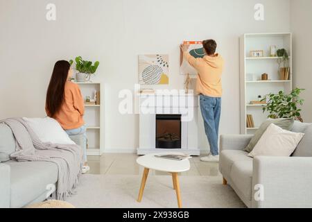 Young man with level hanging painting on wall above fireplace and his wife at home, back view Stock Photo