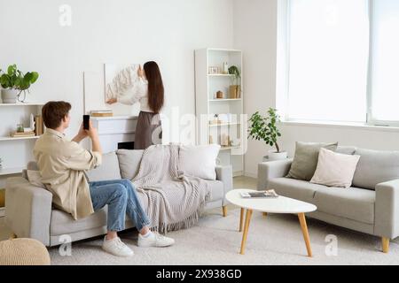 Young woman hanging painting on light wall above fireplace with her husband at home Stock Photo