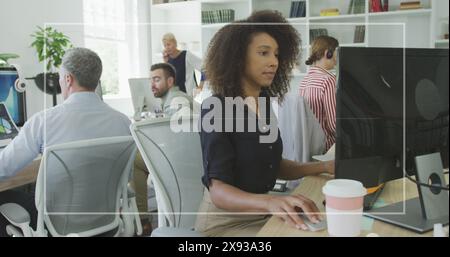 African American woman working on computer in busy office with diverse colleagues Stock Photo