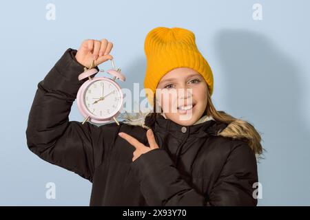 Change to winter time for children. A girl in winter clothes points to an alarm clock with a dial Stock Photo