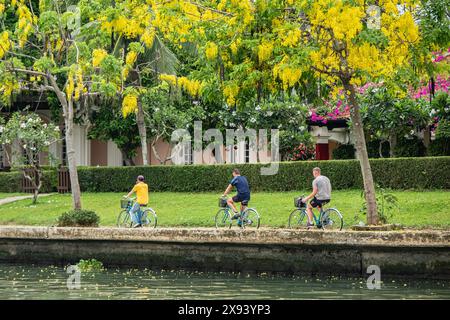 Three man cycle on a path along a river in old town of Hoi An Vietnam. A group of cyclists along an asphalt road by a beautiful river. Bicyclists alon Stock Photo