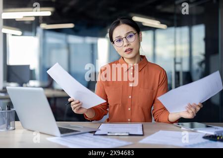 Confused businesswoman wearing glasses reviews documents at her desk in a modern office environment. Stock Photo