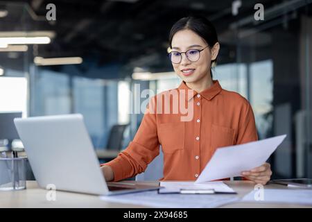 Young professional woman working on a laptop in a modern office setting, holding documents and focused on her tasks. Stock Photo