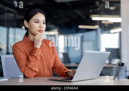 Confident businesswoman deep in thought, working on a laptop at a modern office, embodying focus and determination. Stock Photo