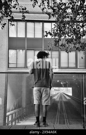 A Young Man standing on High Line Park, NYC, looking out over a Glass Railing with a view of an Image of Train Tracks on a Building's Facade Stock Photo