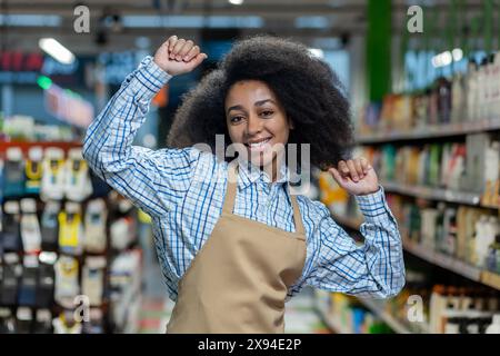 Smiling supermarket employee with afro hair dancing joyfully in the store aisle, wearing an apron and checkered shirt. Stock Photo