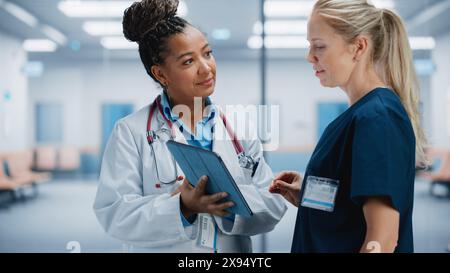 Medical Hospital: Professional Black Female Medical Doctor Talks with White Female Nurse Using Digital Tablet Computer. Two Health Care Specialists Discuss, Find Treatment Solution to Save Lives Stock Photo