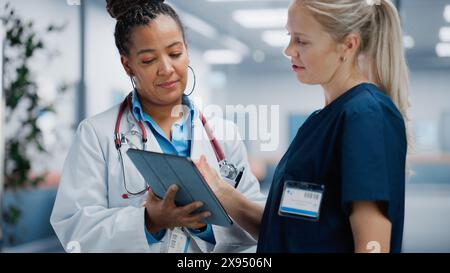 Medical Hospital: Professional Black Female Medical Doctor Talks with White Female Nurse Using Digital Tablet Computer. Two Health Care Specialists Discuss, Find Treatment Solution to Save Lives Stock Photo