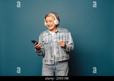 Cheerful, fashionable woman with grey hair dances and smiles in a studio, wearing a denim outfit. Holding a smartphone, she listens to music. Stock Photo