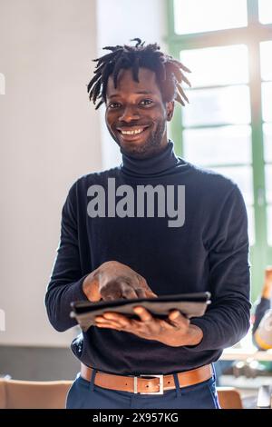 A smiling young African man using a digital tablet indoors. The image captures the essence of modern technology use, showcasing a cheerful, tech-savvy Stock Photo
