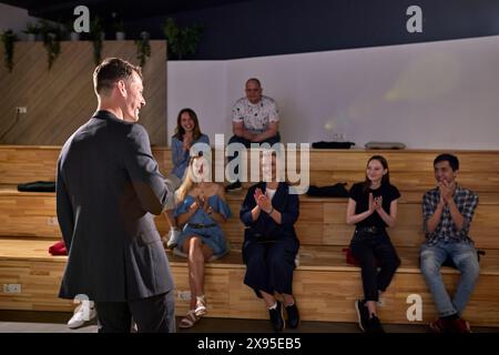 group of young people clapping hands after listening the conference in lecture hall. Rear view on successful caucasian male speaker in formal black su Stock Photo