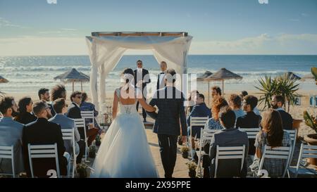 Beautiful Bride in White Wedding Dress Going Down the Aisle with Her Father, while Groom Waits at an Outdoors Ceremony Venue Near the Sea with Happy Multiethnic and Diverse Friends. Stock Photo