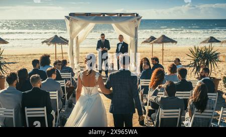 Beautiful Bride in White Wedding Dress Going Down the Aisle with Her Father, while Groom Waits at an Outdoors Ceremony Venue Near the Sea with Happy Multiethnic and Diverse Friends. Stock Photo