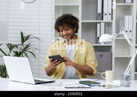 Young man using a calculator for paperwork in a modern home office. He is focused and happy while working on his finances. Stock Photo