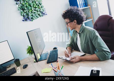 Photo of cheerful handsome agent dressed khaki shirt eyewear writing documents modern gadget indoors workshop workstation Stock Photo