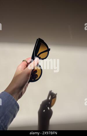 Female hand holding stylish sunglasses over the white wall with a shadow reflecting on it Stock Photo