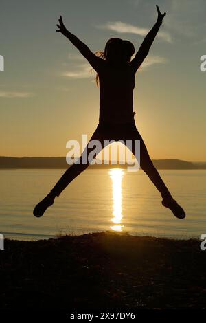 Silhouette of a person jumping joyfully against a sunset over a calm lake Stock Photo