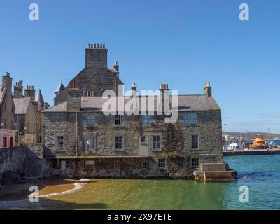 Historic building on the shore overlooking the blue-green sea and a clear sky, grey houses with colourful flags in a seaside town, Lerwick, Scotland Stock Photo