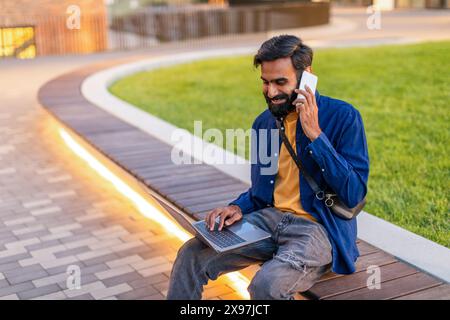 Man Sitting On Bench Using Laptop And Talking On Phone Stock Photo