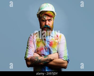Serious professional painter or decorator in splattered shirt and hard hat with crossed arms. Bearded man in protective helmet. Portrait of Stock Photo