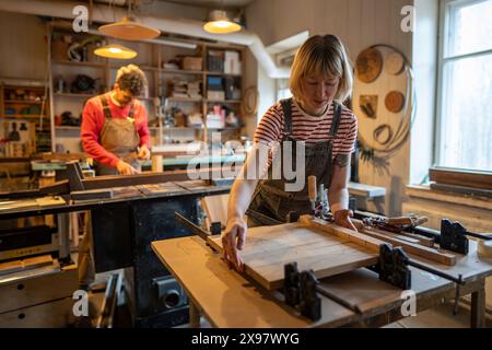 Concentrated female carpenter working on gluing wooden planks together, using bar clamps. Woodwork.  Stock Photo