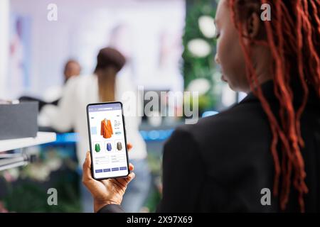 Customer looking at online clothing app shop, buying clothes on website while she sits in fashion boutique at mall. Buyer shopping online on digital store, retail merchandise items. Stock Photo