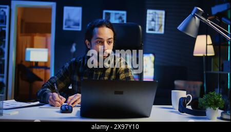 Remote employee working at home office desk, reading information on laptop and writing in notebook. Freelancer noting down ideas on paper using pen while teleworking, camera B Stock Photo