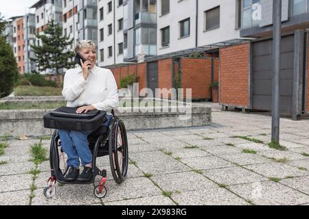 Smiling woman, a wheelchair user with a laptop bag in her lap, using a smartphone to talk. Disability, job, and career concepts. Stock Photo