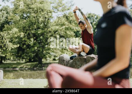 Active friends enjoying a workout session in a sunny urban park, stretching on rocks by a pond Stock Photo