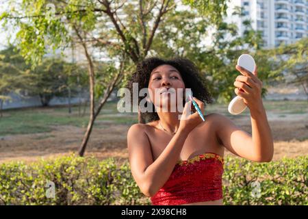 A woman in a red dress applies makeup using a compact mirror in an urban park, surrounded by greenery and modern high-rise buildings, highlighting a m Stock Photo