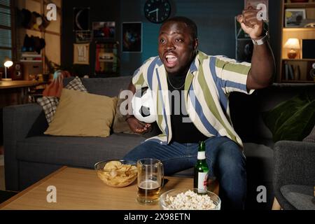 Medium long shot of joyful African American guy holding ball sitting on sofa in living room watching soccer championship match on TV Stock Photo