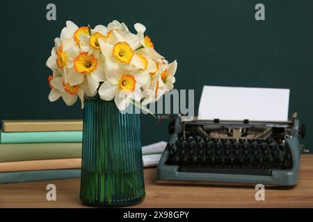 Vase with yellow flowers, retro typewriter and books on wooden table against green background Stock Photo