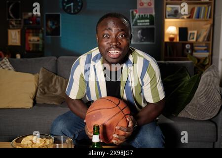 Medium shot of cheerful African American guy sitting on couch watching basketball match on TV late in evening Stock Photo