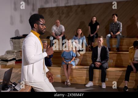Afro lecturer presenting lesson with microphone to caucasian group of people. Young black teacher talking about finance in lecture hall. Side view on Stock Photo