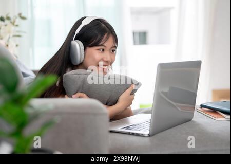 A cheerful young Asian woman wearing headphones, lying down on a sofa in the living room, and enjoying watching a movie on her laptop computer. hobby, Stock Photo