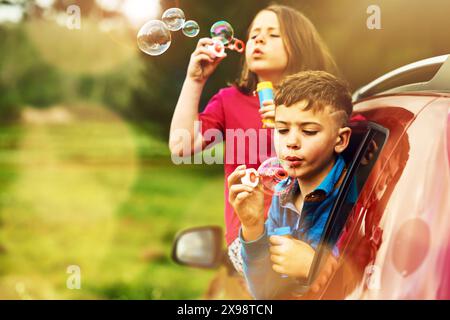 Blowing, bubbles and kids for fun by window in nature, outdoor or countryside with soap toys. children, boy and girl in car for playing, freedom and Stock Photo