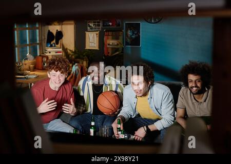 Group of diverse male friends sitting on couch watching basketball match on TV late in evening Stock Photo