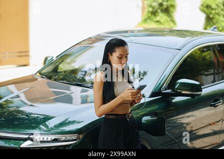 Young businesswoman standing at her electric car and checking notifications on smartphone Stock Photo