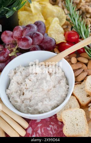 Close-up of a creamy dip in a white ceramic bowl, surrounded by a colorful assortment of appetizers on a charcuterie board Stock Photo