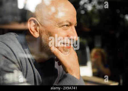 Thoughtful senior man with a beard looking out through a glass window, reflecting on something Stock Photo