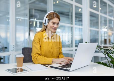 Smiling woman programmer working remotely with laptop and headphones in a modern office. Concept of remote work, technology, and professional development. Stock Photo